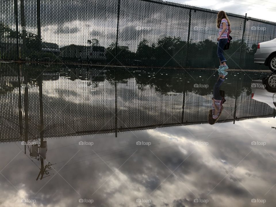 Urban fence and cloudy sky reflected in puddle as girl with blue shoes walks by silver car