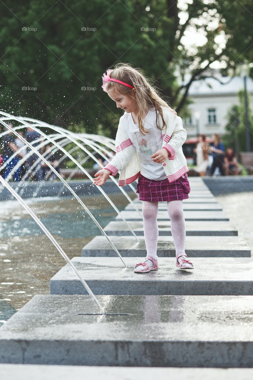 Little happy girl playing with the water in fountain in the center of town. Child catching a water stream going from a fountain. Candid people, real moments, authentic situations