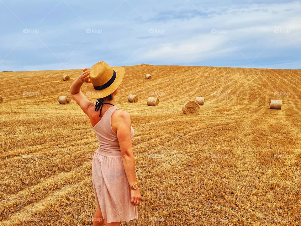 woman from behind with straw hat and one-shoulder summer dress immersed in a golden wheat field