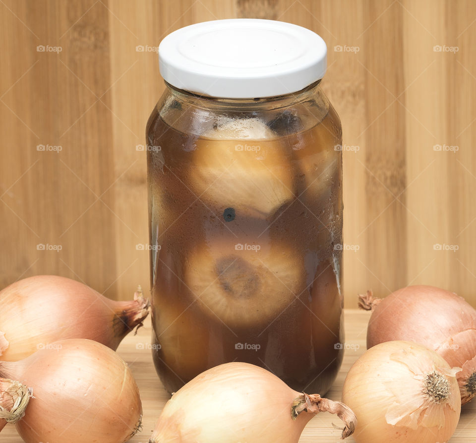 A jar of homemade pickled onions surrounded by small onions in skins against a wooden background.