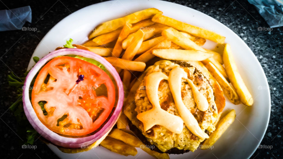 burguer with a top of crab and sauce, tomatoes, onions and lettuce on the side french fries.