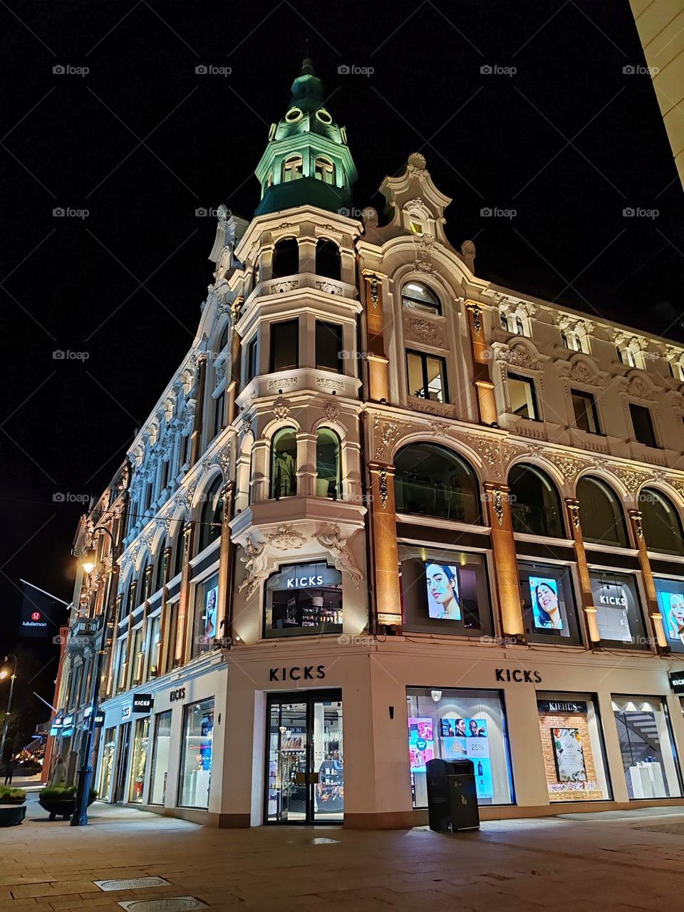 The image shows a large, ornate building at night, illuminated by streetlights and the lights inside the building, which appears to be a store called "KICKS".