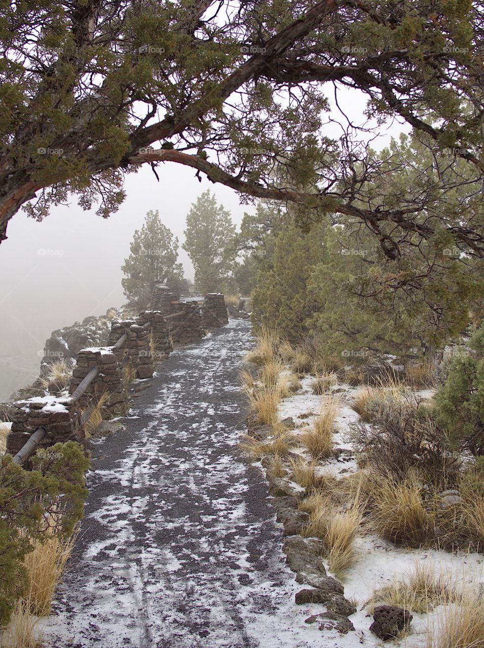 A hiking trail with a scattering of snow at Smith Rock State Park in Central Oregon winds by a cliff with a wooden pole and porous rock foundation railing on a foggy winter afternoon. 