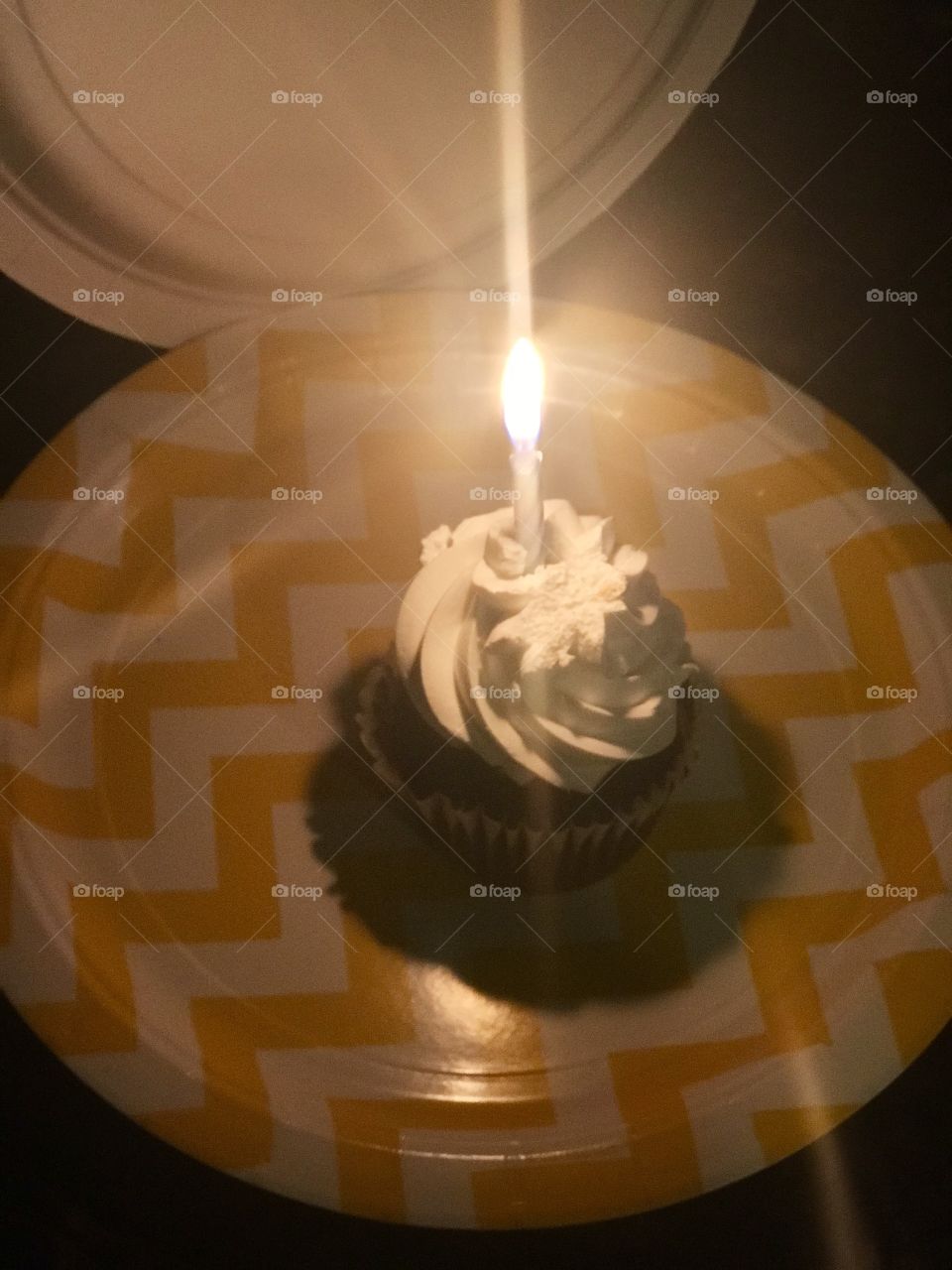A glowing candle Happy birthday chocolate cupcake with vanilla frosting on display on a yellow and white checkered pattern paper plate.