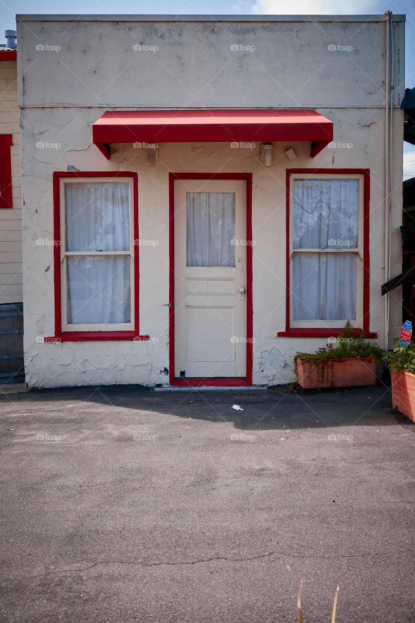 A small cream colored crumbling stucco building with red highlights around the door and windows with a red awning over the door.