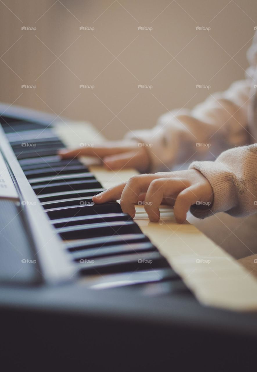 Hands of a little caucasian girl pressing her fingers on the keys of an electric piano while sitting on a sofa in the living room, close-up side view. Music education concept, musical instruments.