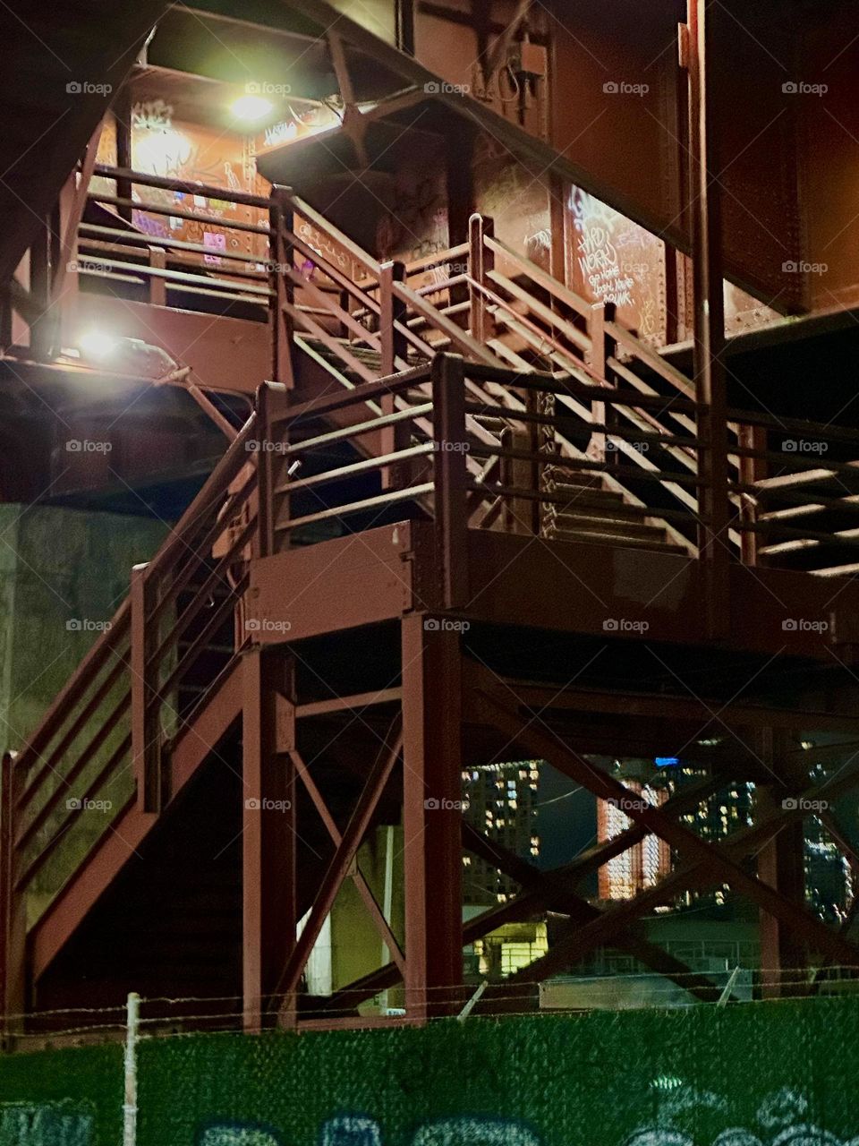This is the red metal staircase of the “Pulaski Bridge” at “Newtown Creek” on the “Greenpoint”, Brooklyn side of the bridge. It is illuminated at night to show people the way up to the pedestrian lane. 2023. Hypnotic Productions