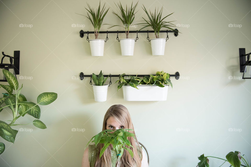 Young millennial woman hiding her face behind a houseplant in a room full of plants