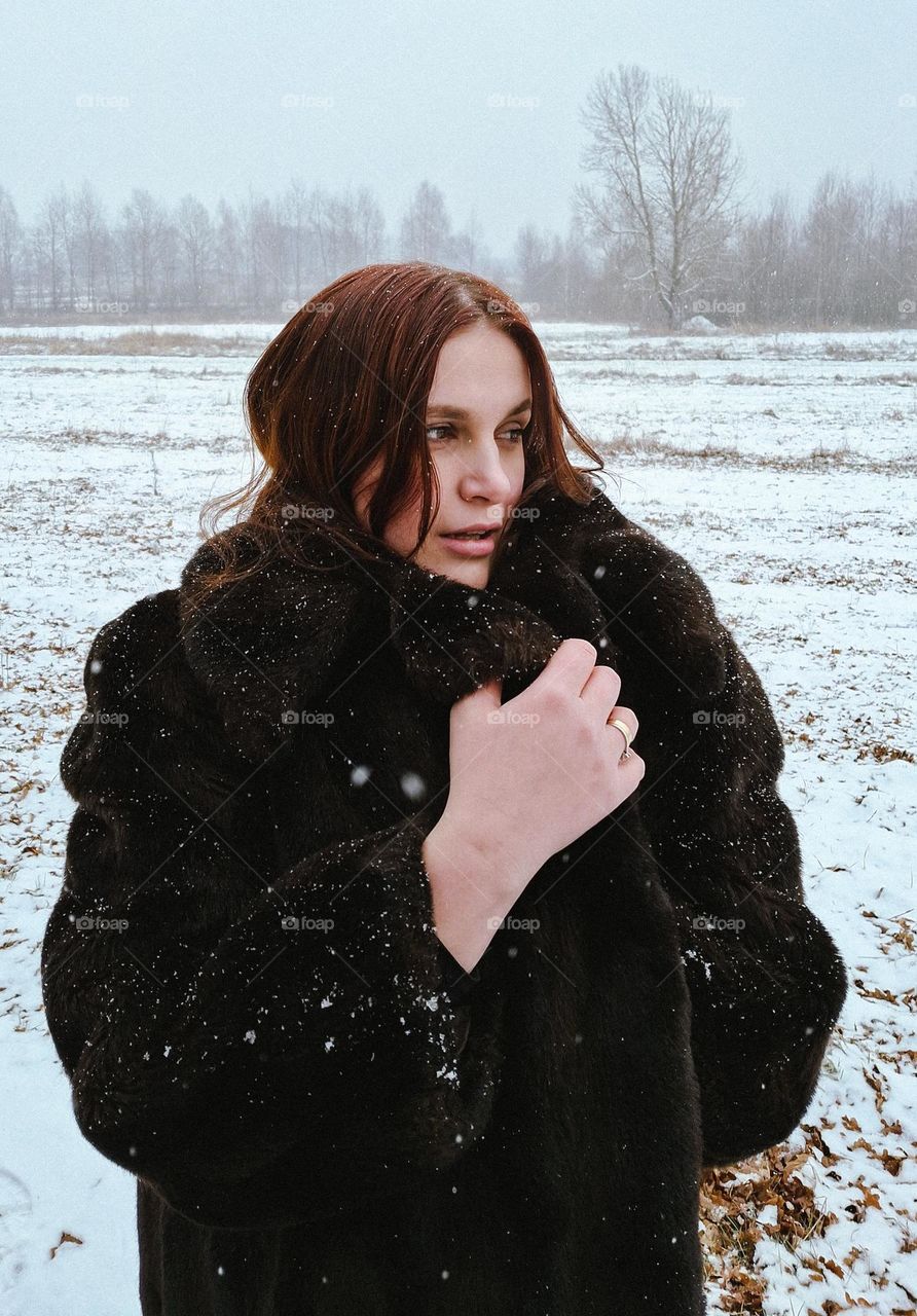 original winter portrait of a light-skinned girl in a dark fur coat amidst snowfall