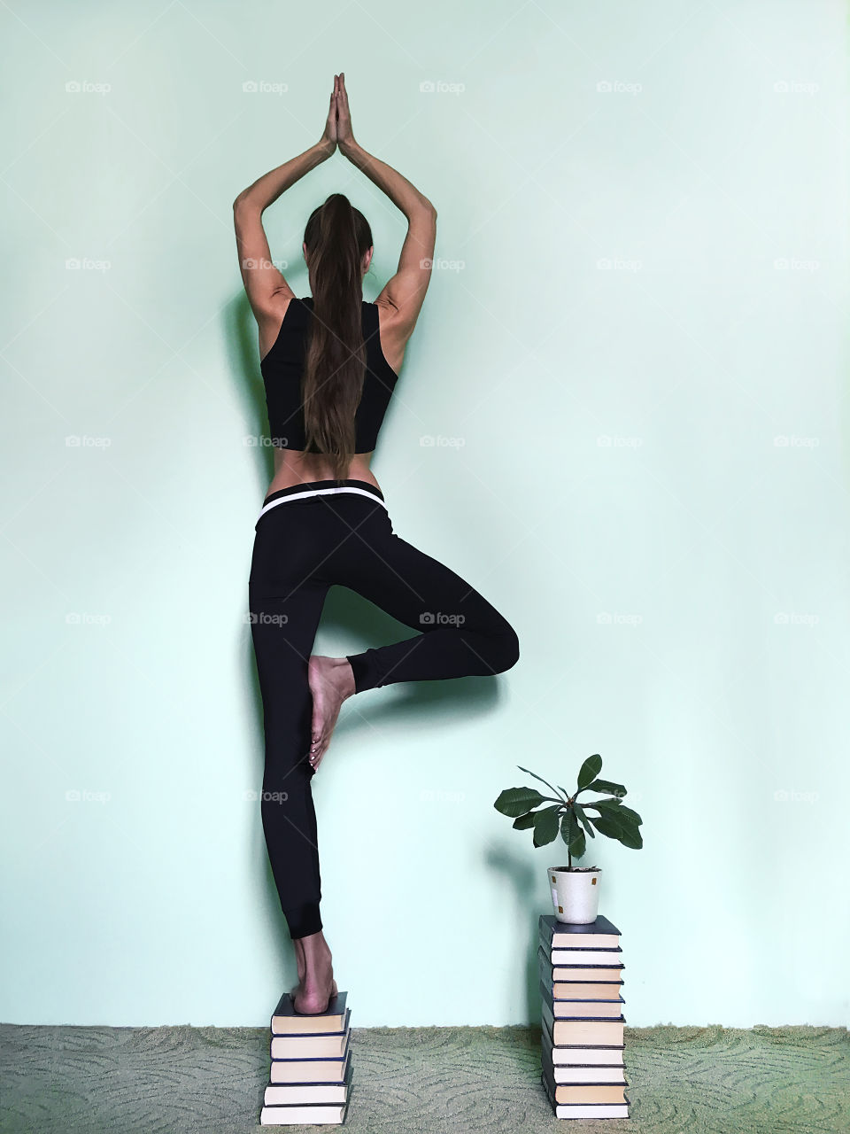 Young woman with long hair doing home yoga and meditating nearby books and house plant on blue monochrome background 