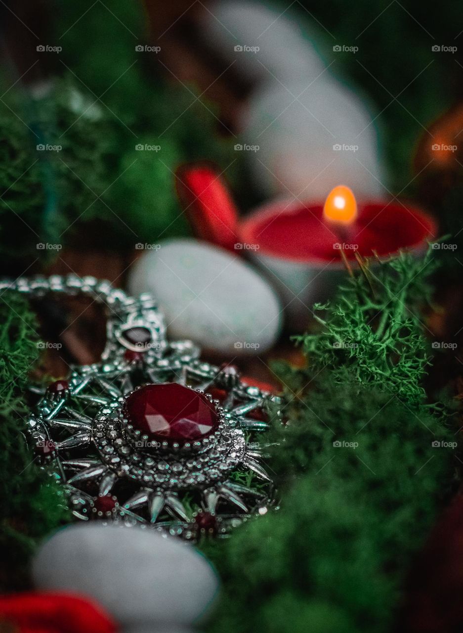 One ancient amulet with red stones on a silver chain lies in a natural tree bark with green nature moss, gray stones, a burning candle and a blurry a jet of smoke on a black background, side view close-up, concept, vintage jewelry, ancient amulet, es