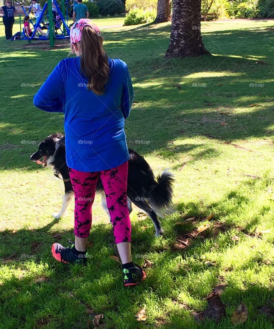 Back view of young girl in bright coloured active wear and baseball cap walking border collie sheepdog on blue leash in grassy park near children's playground 