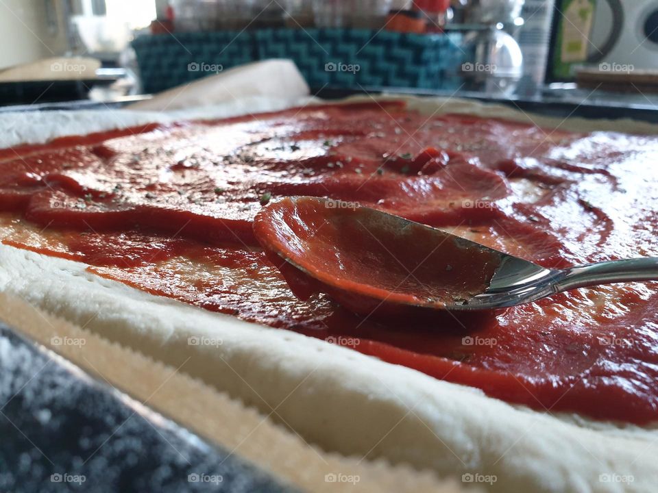 a portrait of a spoon used to spread the red tomato sauce on a pizza dough.