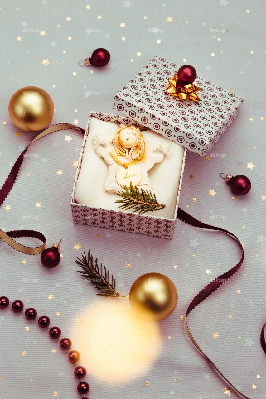 Wrapping Christmas gift into box. Decorating using red ribbon, red Christmas balls and pine twigs. Blurred Christmas lights. Flat top-down composition. Figure of angel made of earthen with love inscription as Christmas decoration