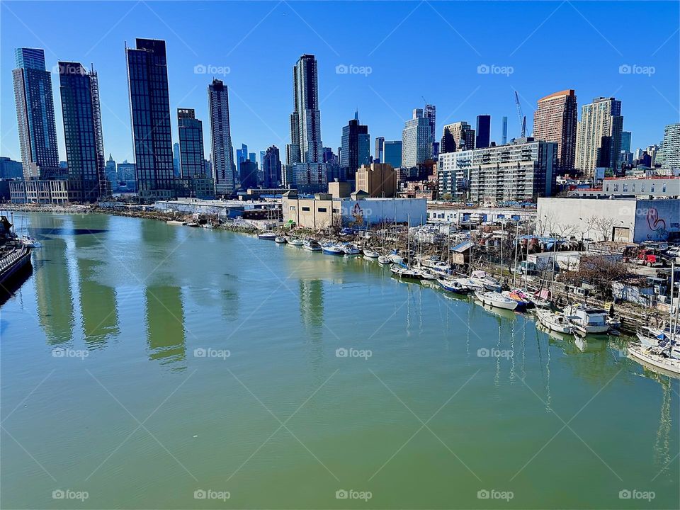 This is beautiful „Newtown Creek“ with its many boats seen from the „Pulaski Bridge“ that connects LIC, Queens to „Greenpoint“, Brooklyn. Across the water behind LIC we see the „Manhattan“ skyline. 2024. Hypnotic Productions