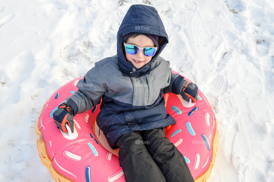 Young boy sitting in a donut innertube and sledding in the snow during winter