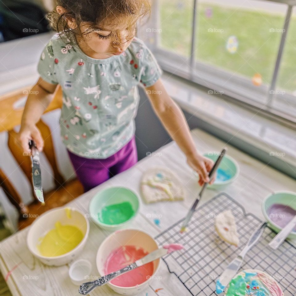 Toddler girl frosting Easter sugar cookies, toddler girl helps mommy in the kitchen, frosting Easter cookies, Easter traditions with family, sweet and delicious iced sugar cookies, baking homemade sugar cookies, little girl decorates Easter cookies