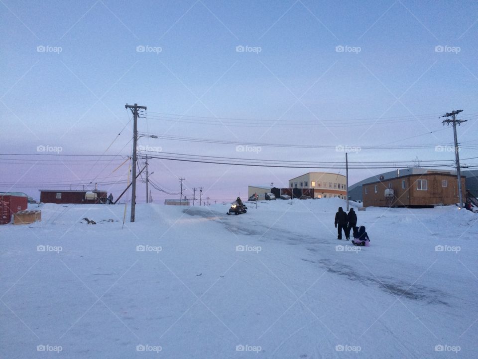 Arctic hamlet winter scene with people towing a sled with a child on it and a person on a snowmobile. 
