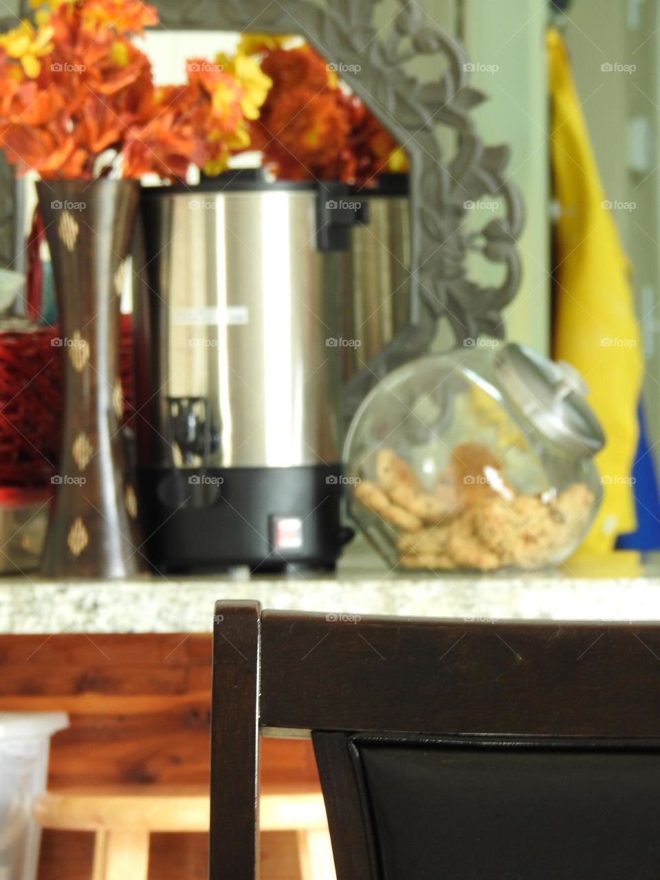 Orange yellow bouquet representing fall autumn behind a woven dark red basket perfect for autumn fall days to warm up with hot chocolate with a large capacity coffee brewer maker and a glass jar with cookies on a kitchen counter and foreground chair.