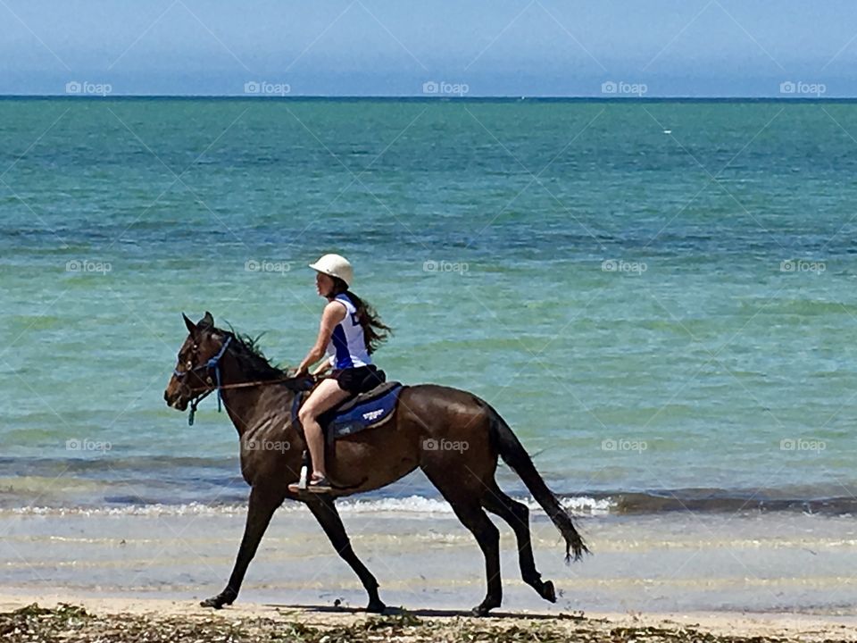 Teenage girl horseback riding cantering along seashore 