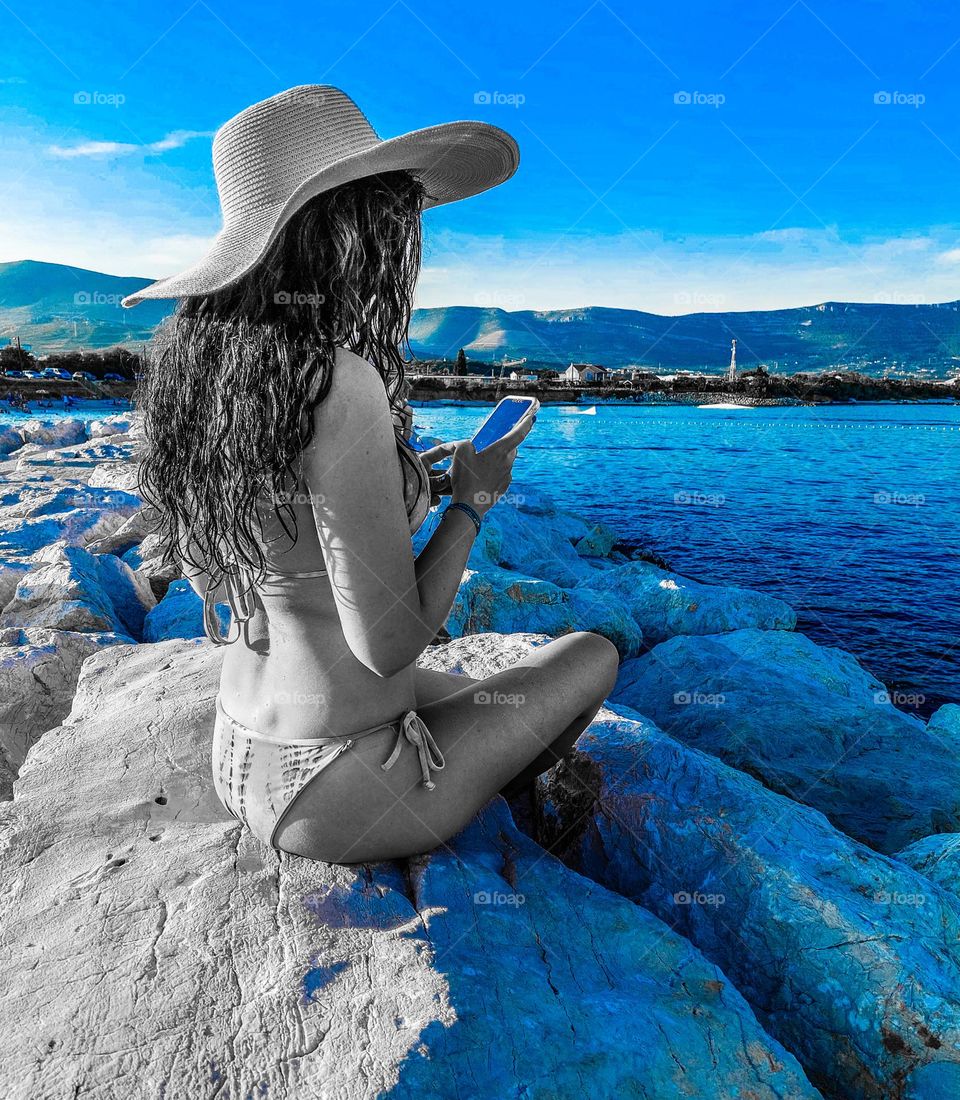 A young woman sits on a rock by the sea and enjoys photographing the seascape with a mobile phone