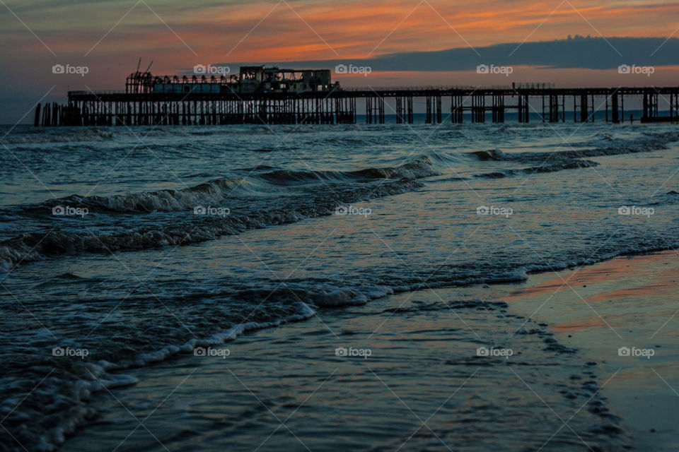 The sea at dusk, waves lap gently on the shore after the sun has gone down, the burnt remains of Hasting pier is silhouetted against the orangey sky (UK 2012)