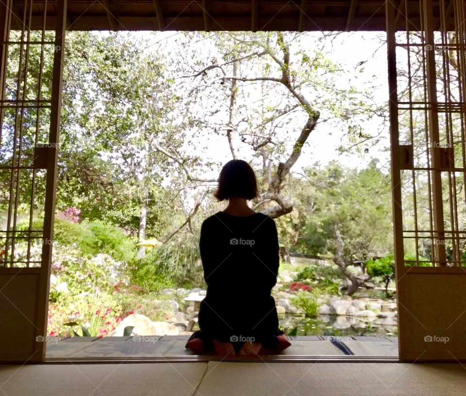 Under gray skies, a woman meditates in a Japanese teahouse overlooking a garden. Storrier Stearns Japanese Garden, Pasadena, California.