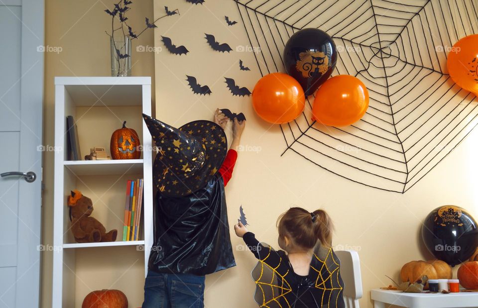 Boy in a black cape and a witch's hat glues a paper bat to the wall. The girl helps her brother, gives him a bat.