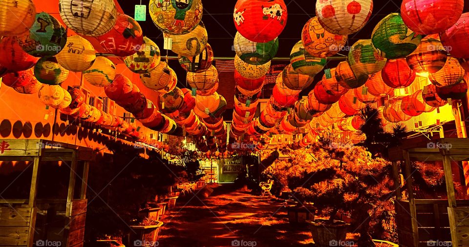 Glowing lanterns hang over a beautiful park. It is nighttime. Some wooden structures are on either side. 