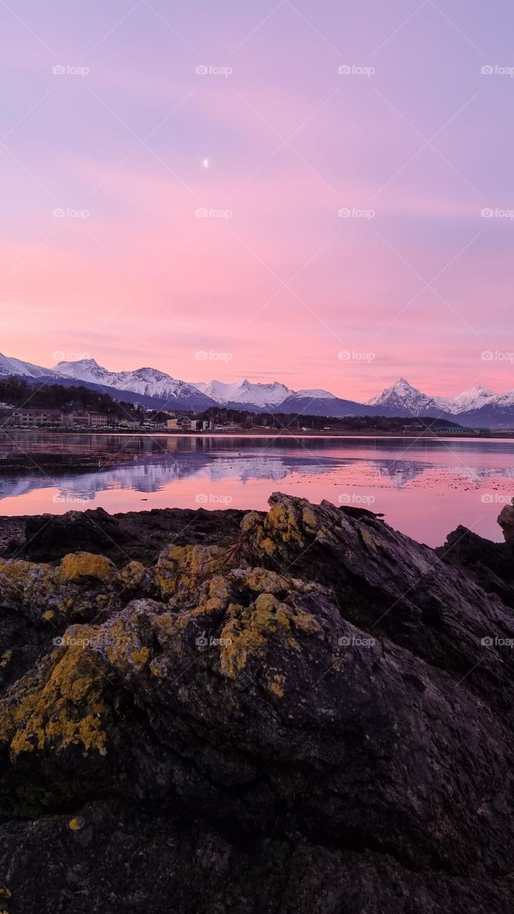 pink sunsetnover all the sky with the moon. Snowy Mountains reflected in the sea water. Rocks.