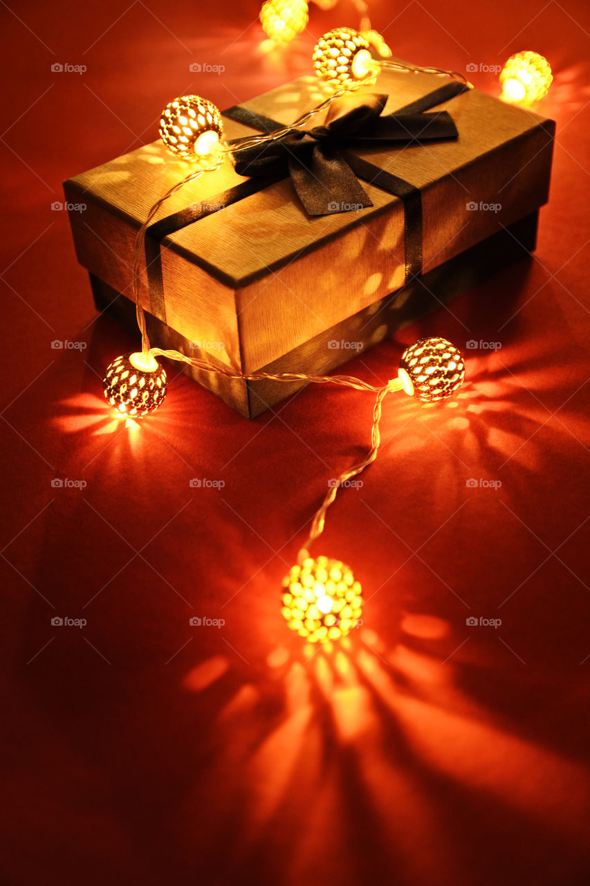 Close up shot of a wrapped gift under glowing Christmas lights on red background, making festive and romantic atmosphere.  Christmas, birthday, anniversary, new year, Valentine's day, date concept.