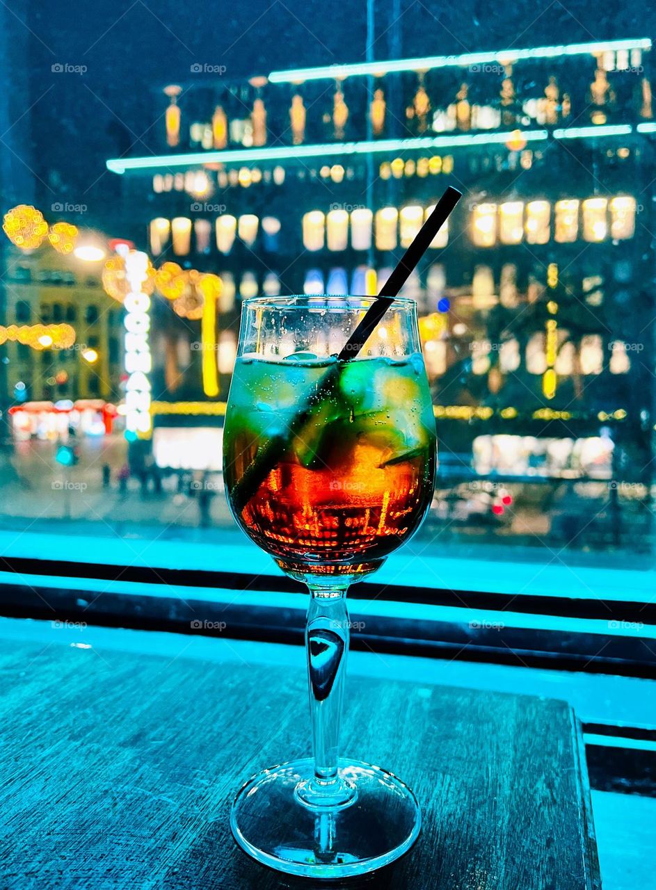 Close view to the drinking glass with tasty cocktail and drinking straw and defocused background cityscape view