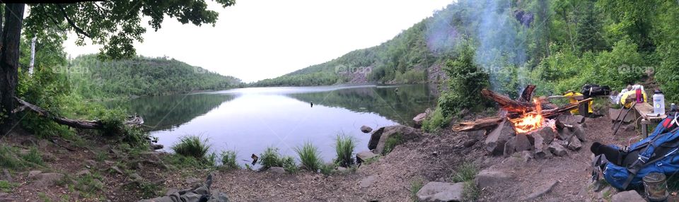 Panoramic shot of a camp scene by the lake that includes a bonfire and a bright view of the campsite