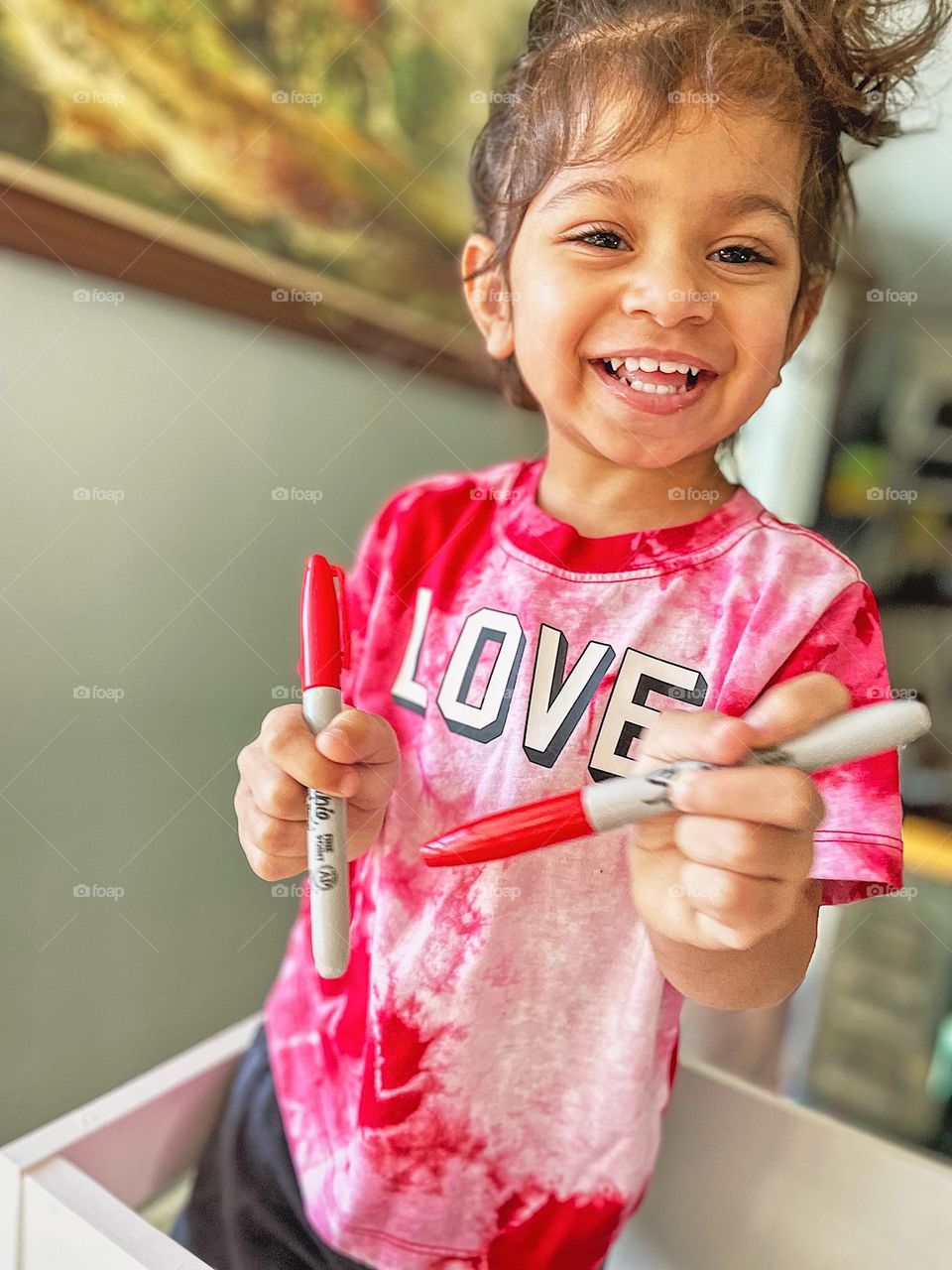 Toddler girl with Sharpie markers in hand, ready to do some crafting, toddler girl smiling in pure joy, toddler gets ready to craft, smartphone photography, portrait of a toddler girl with love t-shirt, love is love 