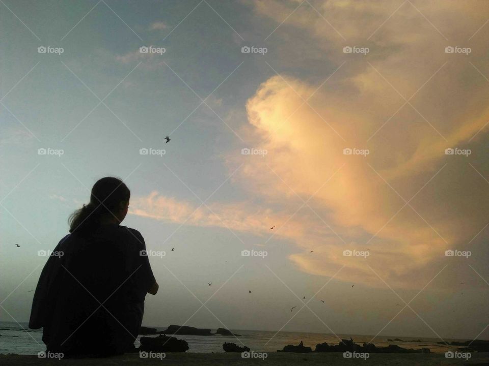 A young girl siiting on wall and looking at the clouds in the sky.