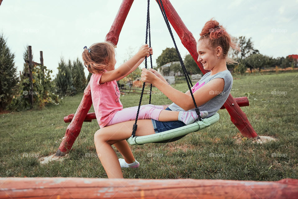 Teenage girl playing with her younger sister in a home playground in a backyard. Happy smiling sisters having fun on a swing together on summer day. Real people, authentic situations