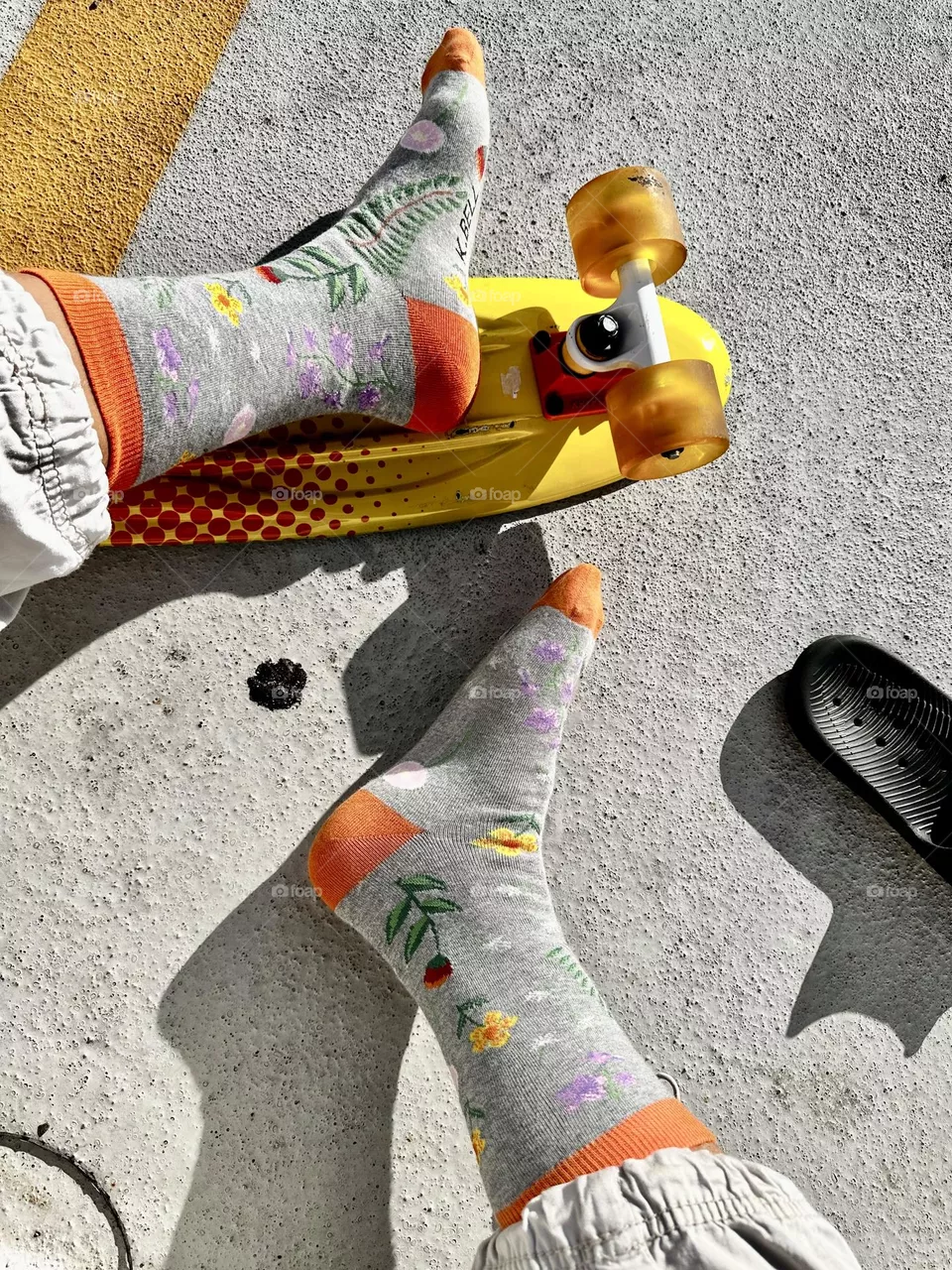 K.BELL grey socks with floral designs and orange top heel and toe, worn with cropped cotton pants, with orange and yellow skateboard in parking lot/skate area