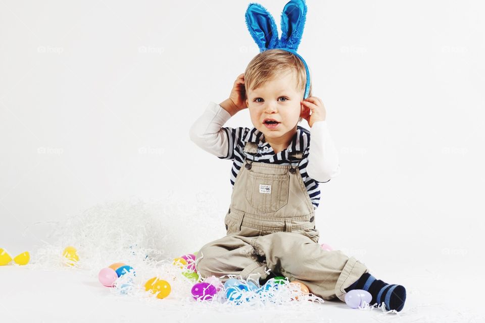 Young boy sitting on whit background with Easter bunny ears and plastic eggs 