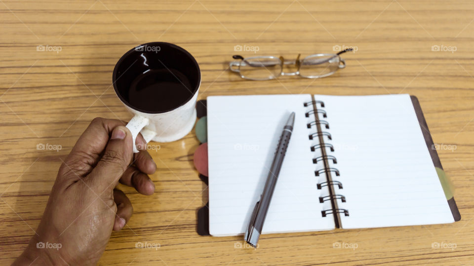 Businessman writing in his diary. Open notebook with blank pages next to cup of coffee and eyeglasses on wooden table. Business still life concept with office stuff on table. Top view with copy space.