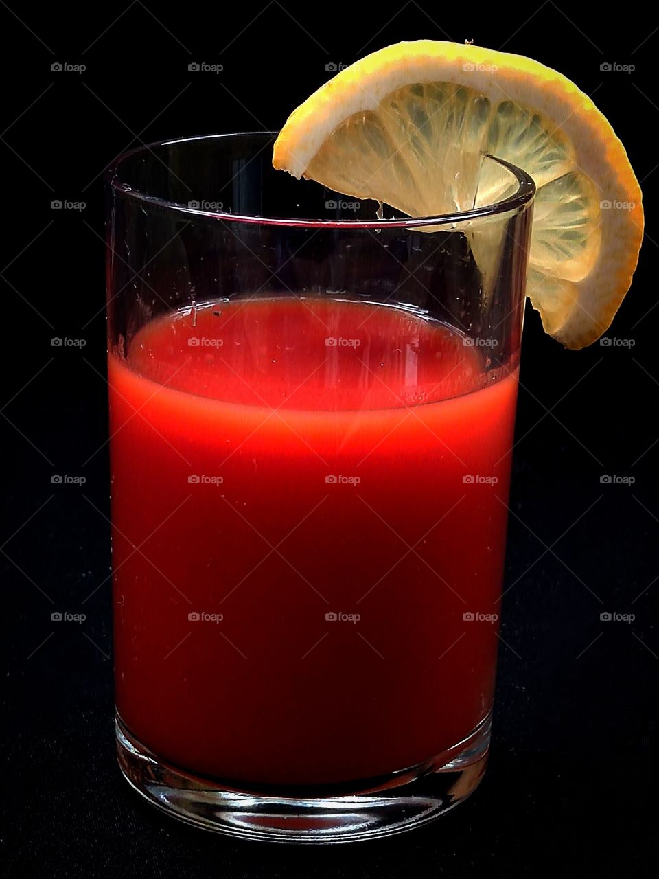 In a glass, red tomato juice. A slice of lemon is suspended on the edge of the glass. Black background