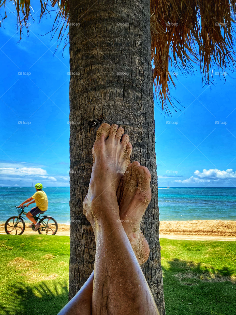 Sandy feet propped up on the trunk of a palm tree as a bicyclist pedals past on the beach bicycle path