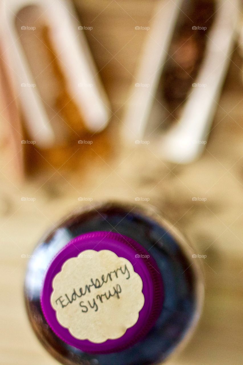 Overhead view of homemade elderberry syrup in a glass bottle with a labeled purple lid, blurred background including whole and ground spices in wooden scoops on a wooden surface 