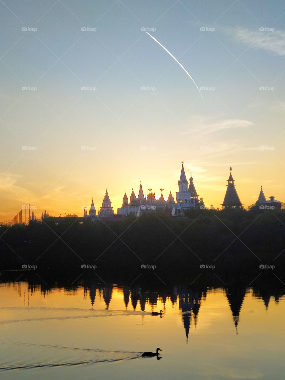 The river is orange from sunset on which ducks swim. The domes of the castle are visible across the river among the green trees. The sun is setting behind one of the domes. The white stripe of the aircraft taking off is visible in the blue sky