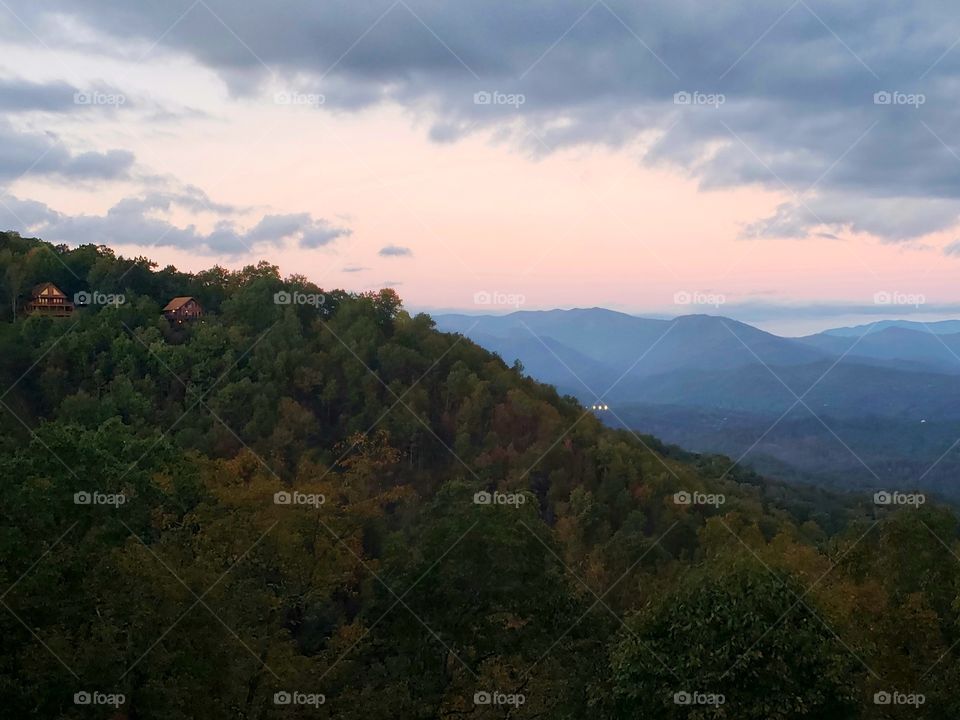 beautiful pink sunset sky above a tree covered mountain with two small cabins near the top and other blue colored mountains in the distance