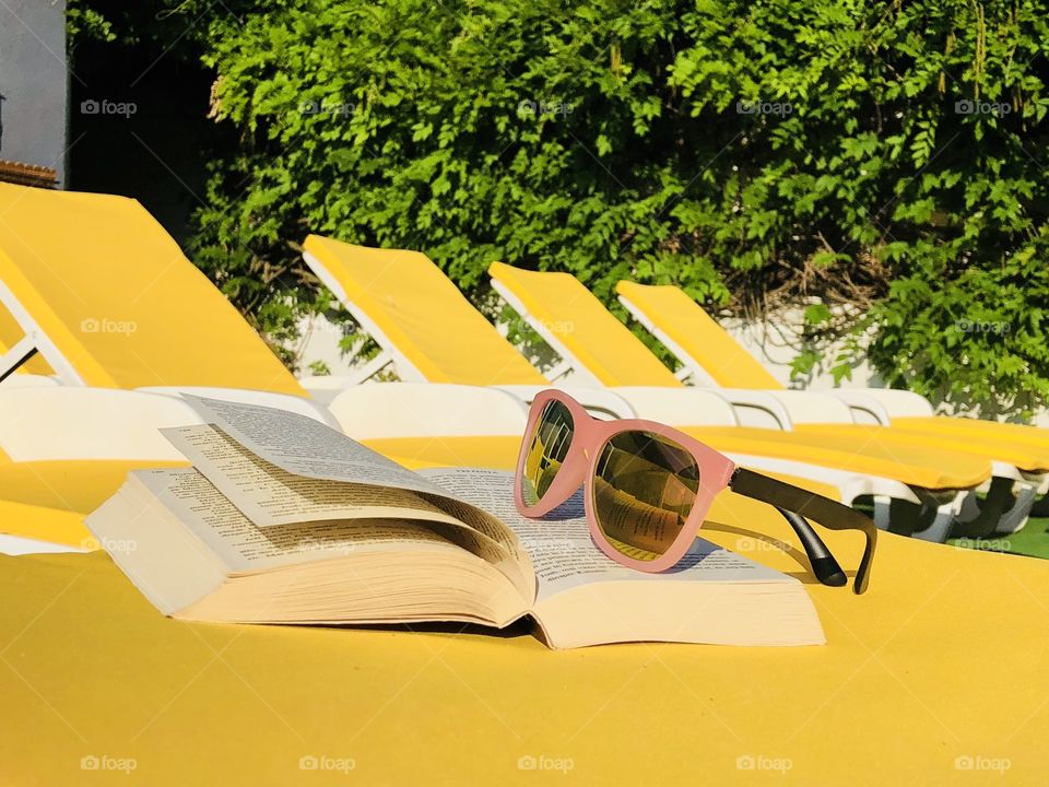 Pink sunglasses and a book placed on a yellow sunbed with many yellow sunbeds in the background