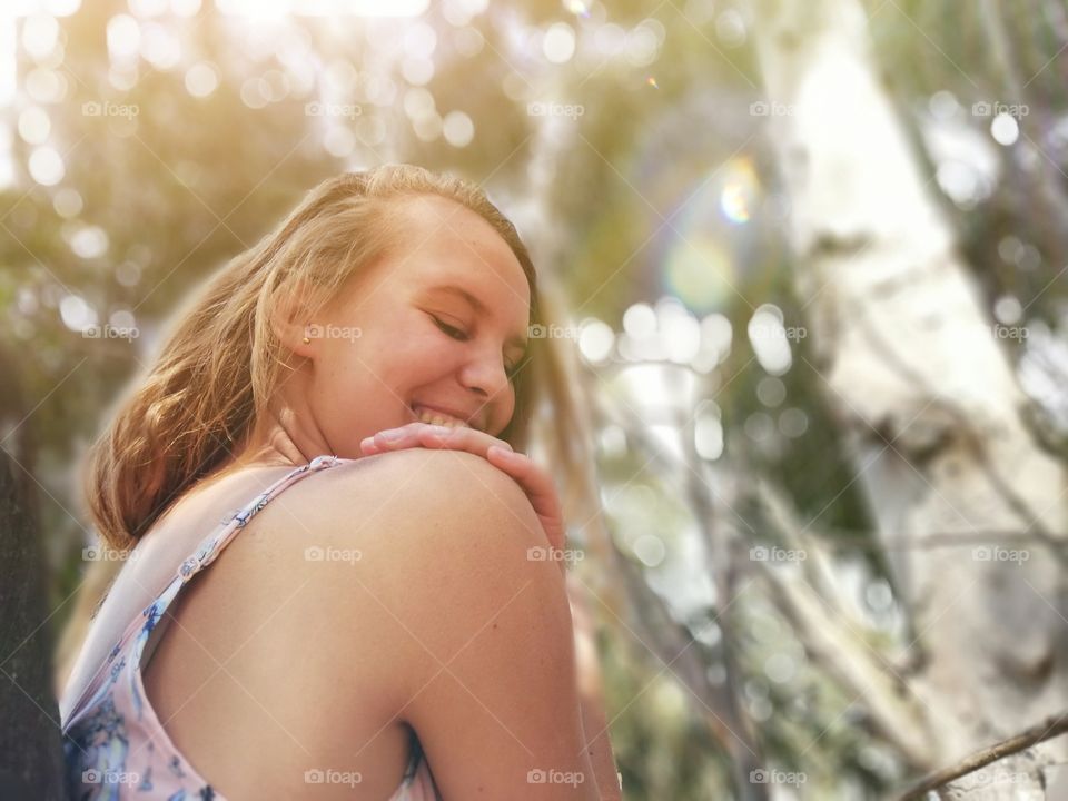 laughing girl sitting under a tree with bokeh's shining in the back ground as the sun shines from behind the Green Leaves