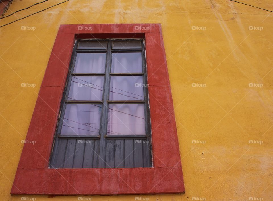 The exterior second floor residential window with a colorful red frame surrounded by the building's yellow concrete wall in San Miguel de Allende, Mexico.
