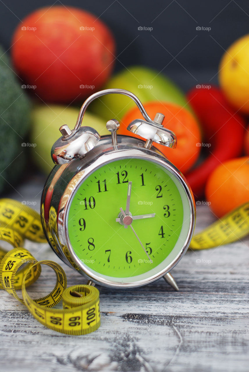Time to loose weight. Green clock with measurring metter and healthy fruits vegetables on background.