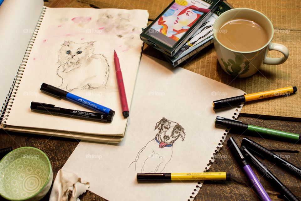 Favorite things: drawing cat and dog pet portraits at home with favorite Faber-Castell PITT Artist pens high angle view of sketches on sketch book paper on wood table with coffee, water for watercolor technique and blotter 