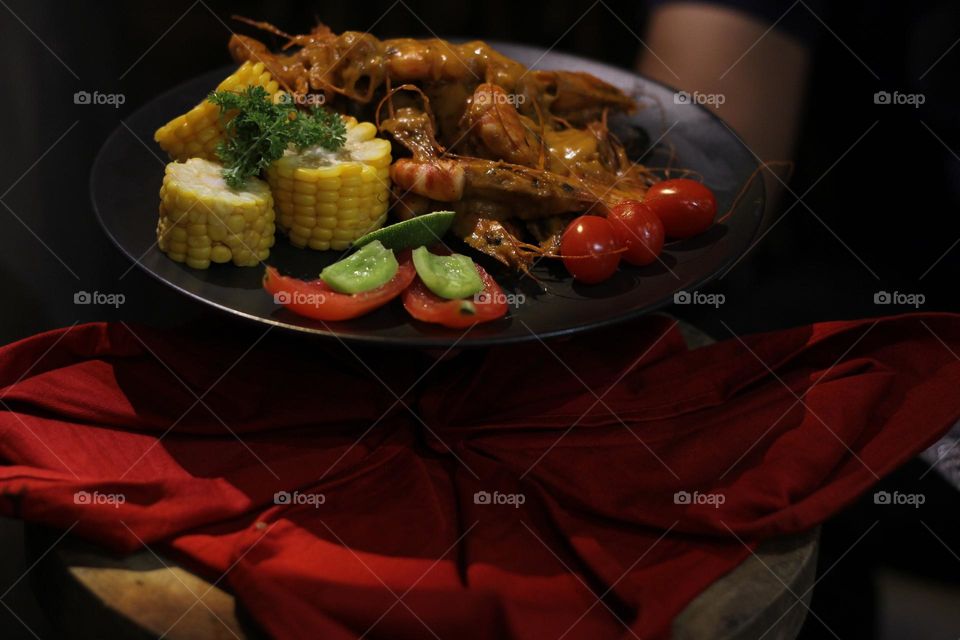 Asian food balado fried prawns with corn chunks, lemon slices and tomatoes served on black plate with red cloth bottom and wooden coaster. Asian style street seafood.